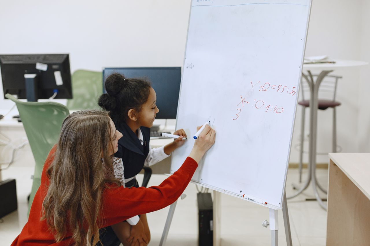 A teacher helps a student with math on a whiteboard in a classroom setting.