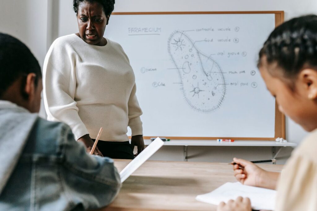 Crop African American female teacher standing near desk while schoolchildren writing information in copybooks during lesson